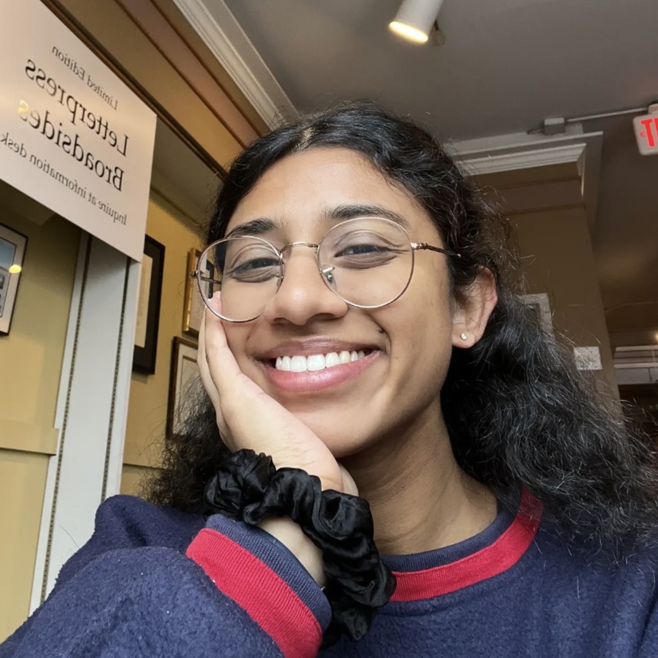 A photo of Amritha with her chin resting in one hand, smiling at the camera. There is a sign behind her, but it is unclear what building she is in. Her hair is half-up, half-down and she is wearing a navy blue sweater with stripes of red at the cuffs and neckline.