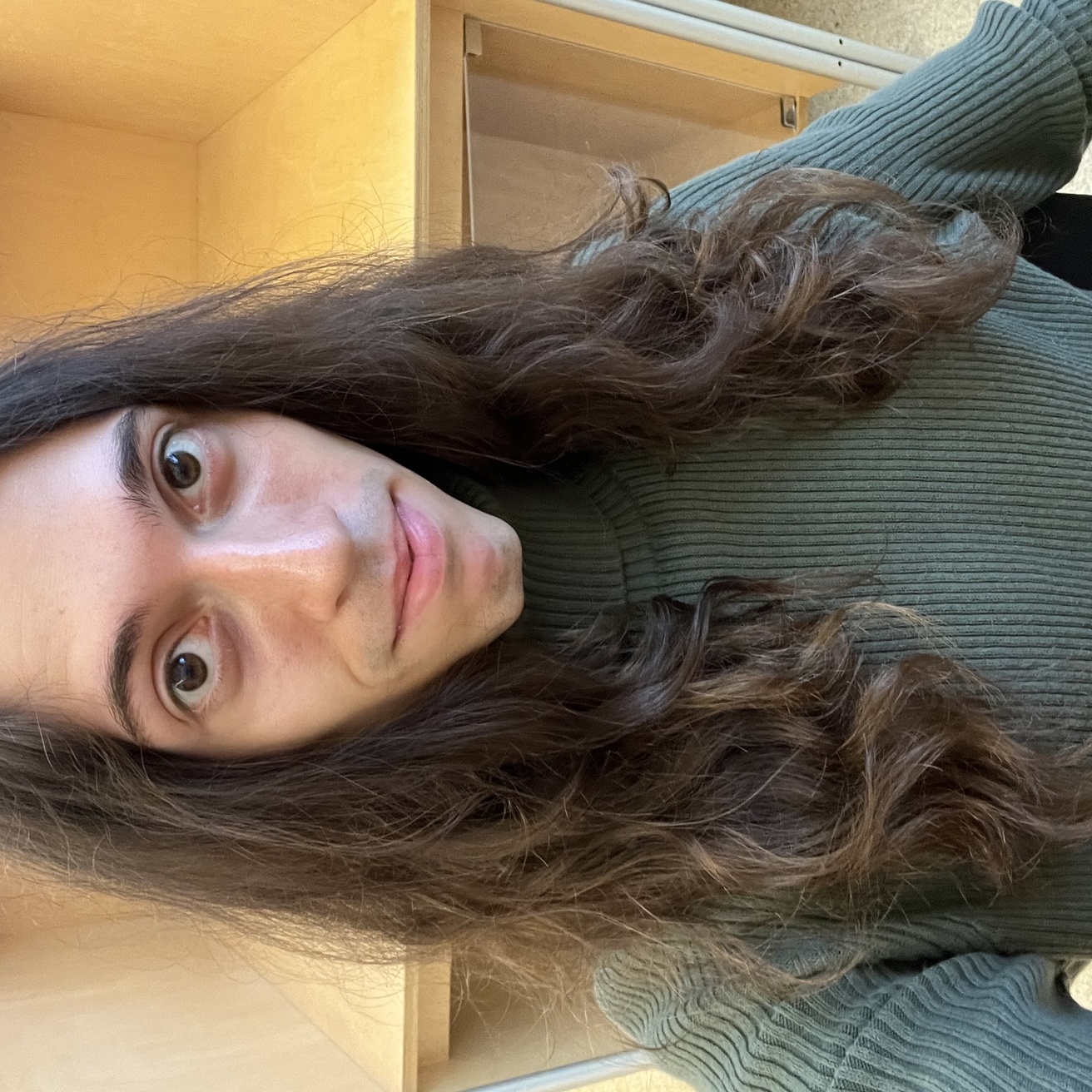 An image of Wyeth standing in front of a bookshelf. His hair is down, hanging in loose curls over his shoulders. He is wearing a dark green turtleneck sweater. 
