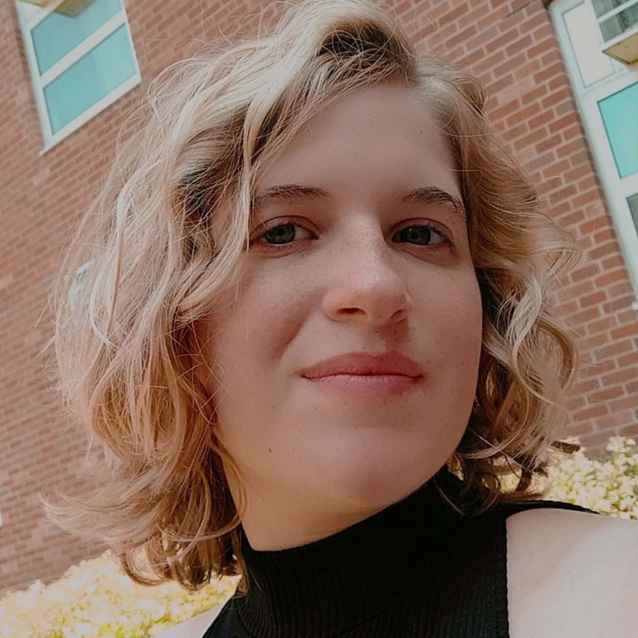 A headshot of Dennie, smiling softly at the camera. They are posed in front of a brick building, with their hair down to their shoulders. They are wearing a sleeveless black turtleneck.