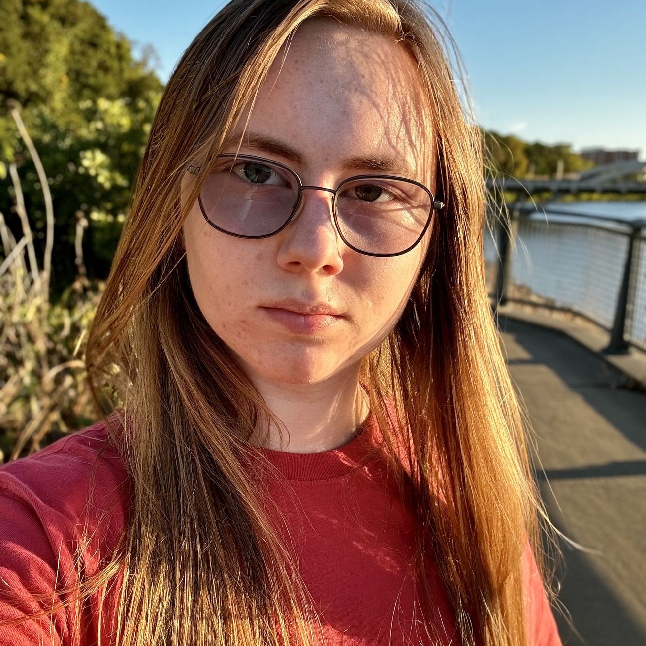 A picture of Noa standing out by the Iowa River near sunset. Their hair is down, falling over their shoulders, and they are wearing a red t-shirt and sunglasses.